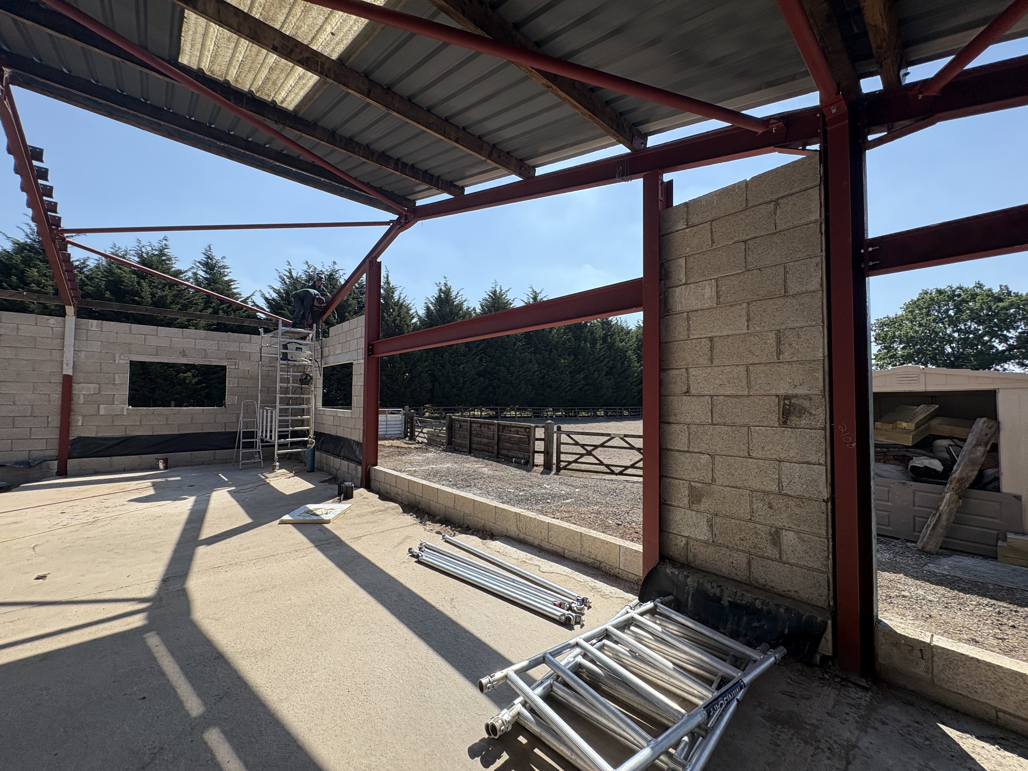 Gable-end view during construction showing new blockwork walls set within the original agricultural steel frame, looking out to the paddock